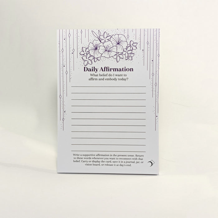 A daily affirmation notepad with a white background, featuring purple floral illustrations at the top, decorative vertical line borders, and lined writing sections in the center. The card is photographed standing upright against a clean white background.