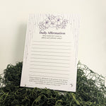 A daily affirmation notepad with floral line art and writing prompts, displayed upright on green moss against a plain white background.