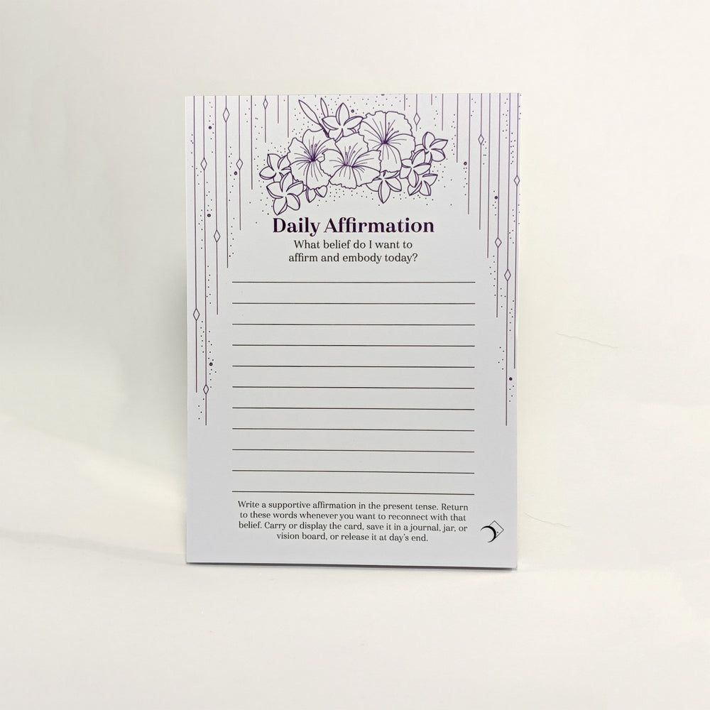 A daily affirmation notepad with a white background, featuring purple floral illustrations at the top, decorative vertical line borders, and lined writing sections in the center. The card is photographed standing upright against a clean white background.