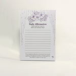 A daily affirmation notepad with a white background, featuring purple floral illustrations at the top, decorative vertical line borders, and lined writing sections in the center. The card is photographed standing upright against a clean white background.