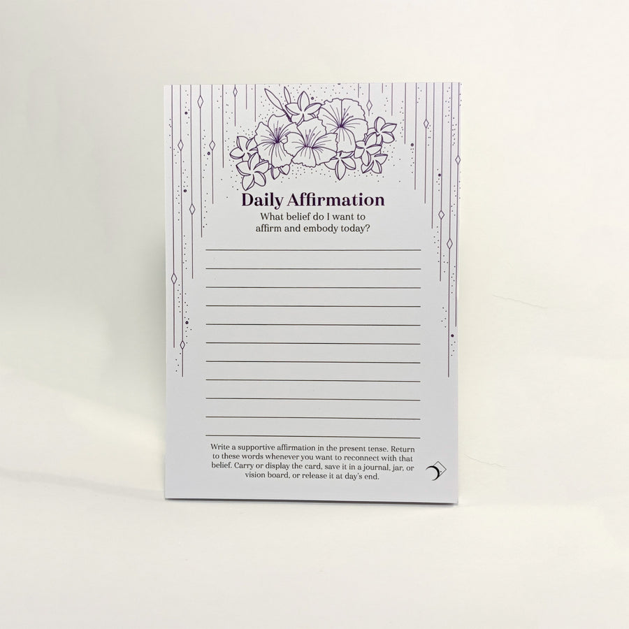 A daily affirmation notepad with a white background, featuring purple floral illustrations at the top, decorative vertical line borders, and lined writing sections in the center. The card is photographed standing upright against a clean white background.