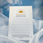 A Daily Intuition notepad with gold decorative lines and text, resting on white draped fabric against a blue sky and clouds background.
