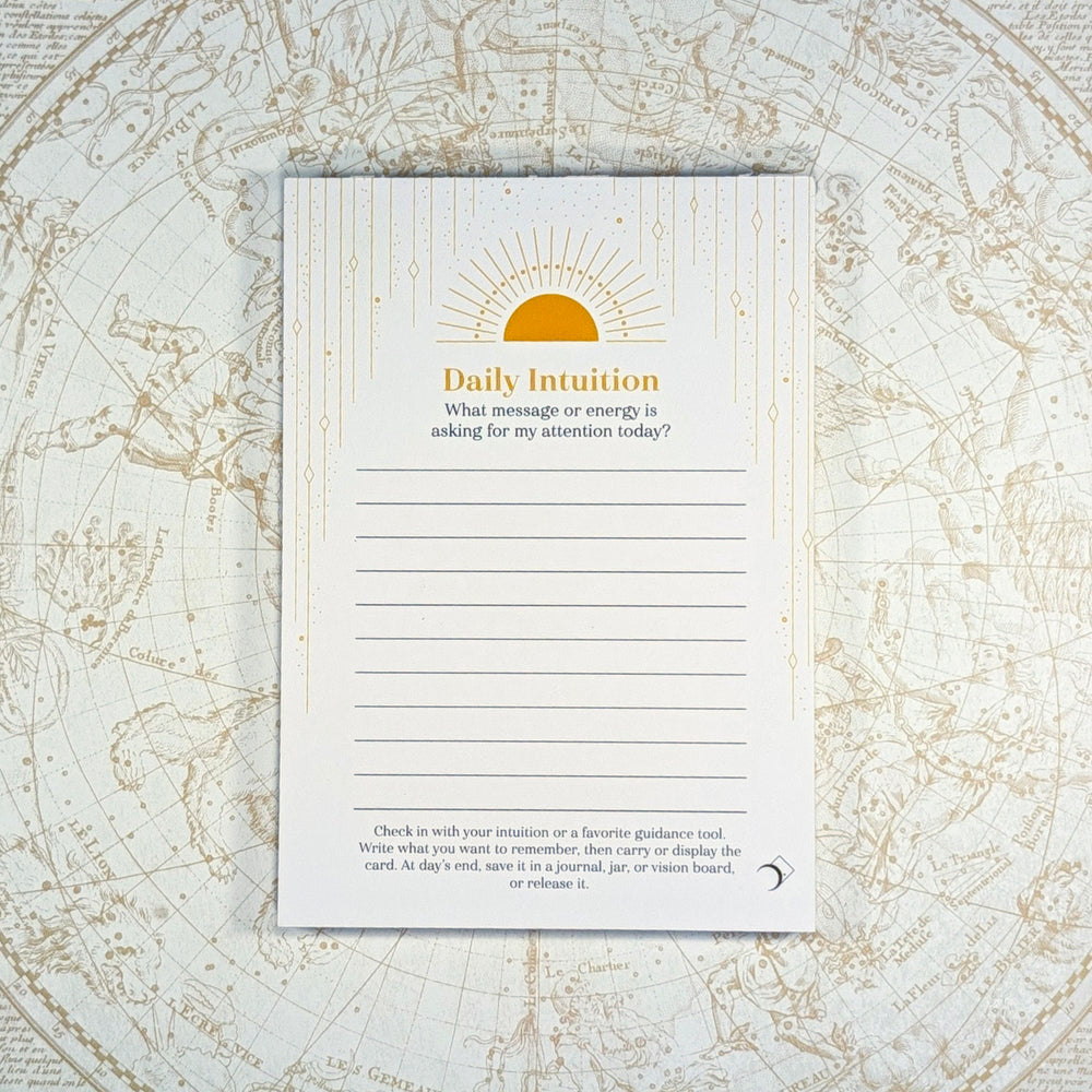 A daily intuition journaling notepad with lined writing space and a gold sun illustration, placed on top of an antique-style celestial map background.