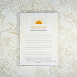 A daily intuition journaling notepad with lined writing space and a gold sun illustration, placed on top of an antique-style celestial map background.