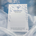 A white Daily Reflection notepad with blue printed shooting star-and-moon illustrations, resting on draped white fabric against a soft blue sky background.