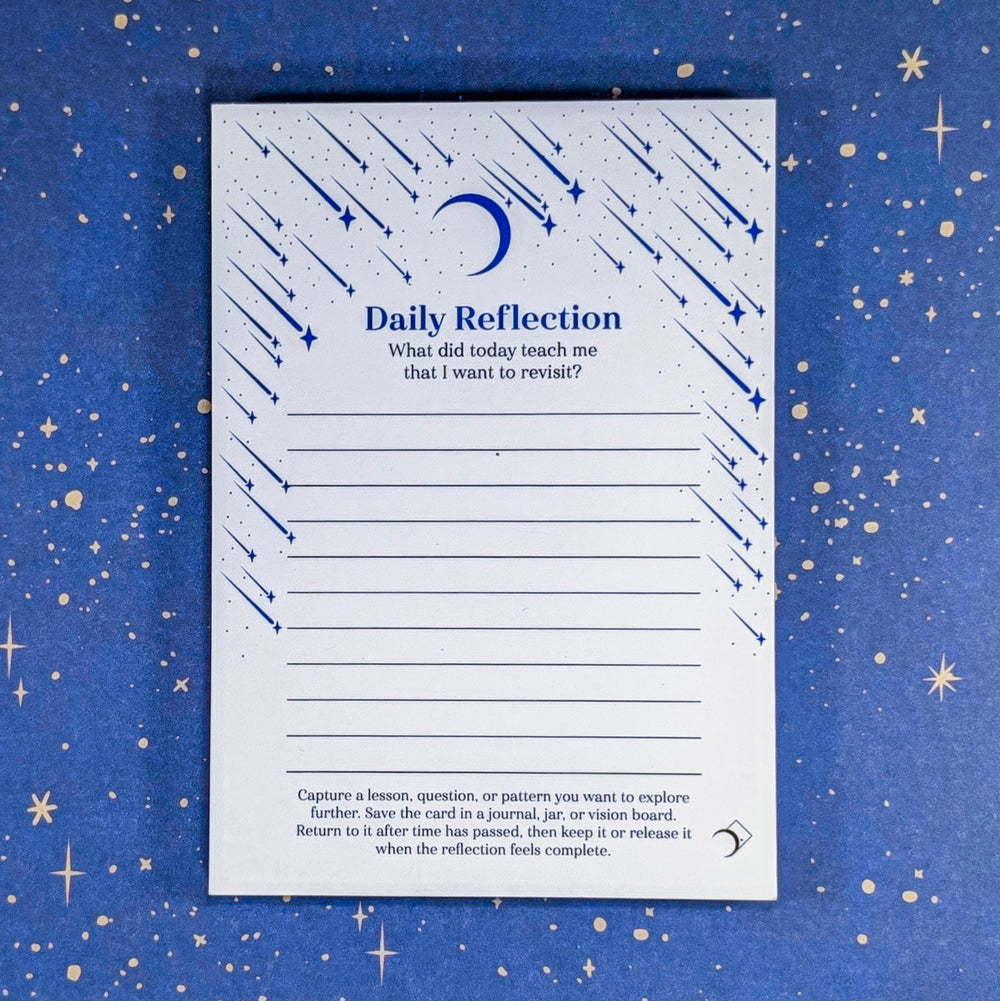 A white Daily Reflection notepad with blue text header black text prompt. Blue shooting stars and a crescent moon illustrations on the notepad. Product laid flat on a blue starry background.