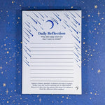 A white Daily Reflection notepad with blue text header black text prompt. Blue shooting stars and a crescent moon illustrations on the notepad. Product laid flat on a blue starry background.