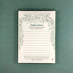 A daily vision notepad with a green floral border and text prompts, photographed flat against a textured green background.