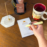 A hand with black and white patterned nail polish holds a yellow pen over a lined daily intention notepad on a wooden table. The scene includes a white marble incense holder with a burning incense stick, a black card holder with oracle cards, and a red-and-white Washington, DC mug.