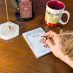 A hand with dark glittery nail polish writes in a lined Daily Vision notepad on a wooden desk, surrounded by a coffee mug, a brass candle holder, and a crystal dish.