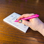 A hand with a gold and black nail design holds a bright pink pen with the words 'WISH TO NEVER GROWN UP' while writing on a white notepad titled 'Daily Reflection'. The scene is set on a warm, polished wooden tabletop.