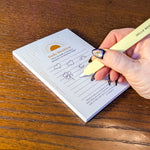 A hand with glittery black nail polish writes in a small white notepad on a wooden table, holding a yellow-colored pen. The notepad has a gold sun illustration and the text 'Daily intention'.