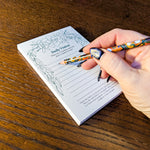 A hand with black and blue glitter nail polish writes in a lined daily vision notepad on a dark wooden table, holding a colorful patterned pencil.